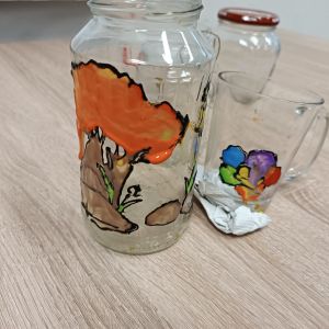 Na stole stoi słoik i szklanka. Na słoiku namalowane jest drzewo, a na szklance kolorowy kwiatek.