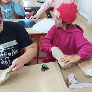 Dwóch uczniów siedzących w ławce trzymających w dłoniach zapakowane w papier pakowny przedmioty.
