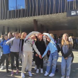 Grupa uczniów stojąca przed wejściem do Muzeum Kinematografii. Chłopiec i dziewczynka układają z rąk serce.