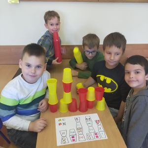 Pięciu chłopców za pomocą żółto czerwonych kubków odwzorowuje obrazek znajdujący się na kartce.