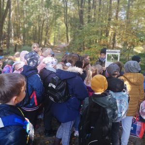 Grupa uczniów stojąca tyłem do obiektywu, słuchająca przewodniczki trzymającej w dłoniach zdjęcie wilka.