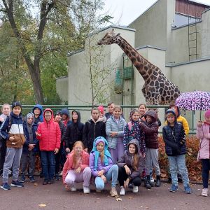 Grupa uczniów wraz z wychowawczynią stojąca przed wybiegiem dla żyraf.