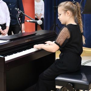 Dziewczynka w czarnej sukience z zaplecionymi w warkocz włosami grająca na pianinie.