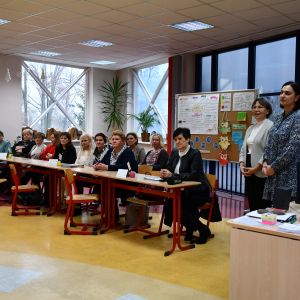 Otwórz zdjęcie - grupa nauczycieli siedzących w ławkach, dwie kobiety stoją obok.