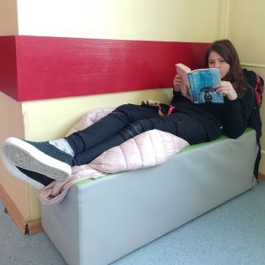 Otwórz zdjęcie -  Uczennica leżąca na pufie stojącej pod ścianą i czytająca książkę.