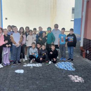 Otwórz zdjęcie - Uczniowie podczas wiosennych zabaw na placu przy szkole rysują kredą chmury.