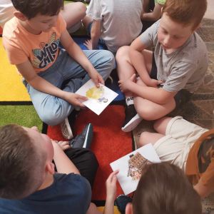 Otwórz zdjęcie -  Uczniowie z klasy 1B siedzący na dywanie próbują dopasować rekwizyt do książki.
