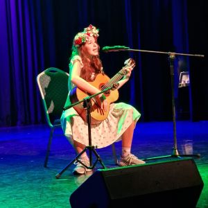 Otwórz zdjęcie - Ania w wianku na głowie siedzi na krześle, gra na gitarze i śpiewa piosenkę o zdrowiu.