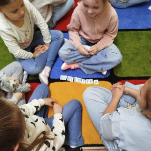 Otwórz zdjęcie  - Dziewczynki układają na dywanie słowo "BIBLIOTEKA".