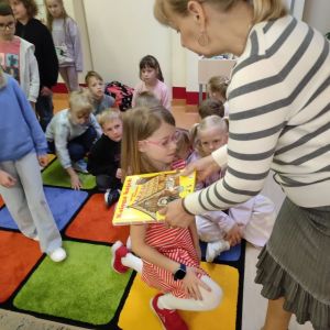 Otwórz zdjęcie - Bibliotekarka kładąc książkę na ramieniu dziecka pasuje je na czytelnika.