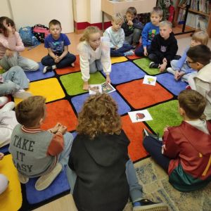 Otwórz zdjęcie - Uczniowie siedzący na dywanie w kole grają w grę zaproponowaną przez bibliotekarki.