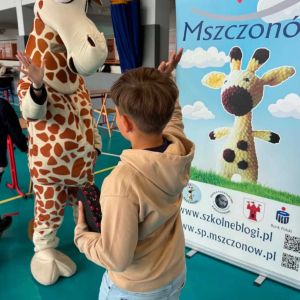 Otwórz zdjęcie - Chłopiec przybija piątkę z dużą maskotką żyrafy obok baneru reklamowego SKO Mszczonów.