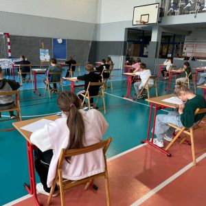 Otwórz zdjęcie - Dzieci siedzą przy osobnych stolikach na sali gimnastycznej i piszą test wiedzy.