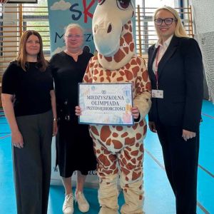 Otwórz zdjęcie - Trzy kobiety i maskotka żyrafy trzymająca plakat olimpiady stoją na hali sportowej.