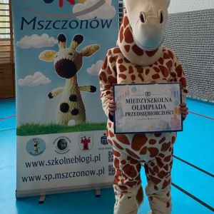 Otwórz zdjęcie - Duża maskotka żyrafy trzyma plakat olimpiady obok stojaka reklamowego SKO.