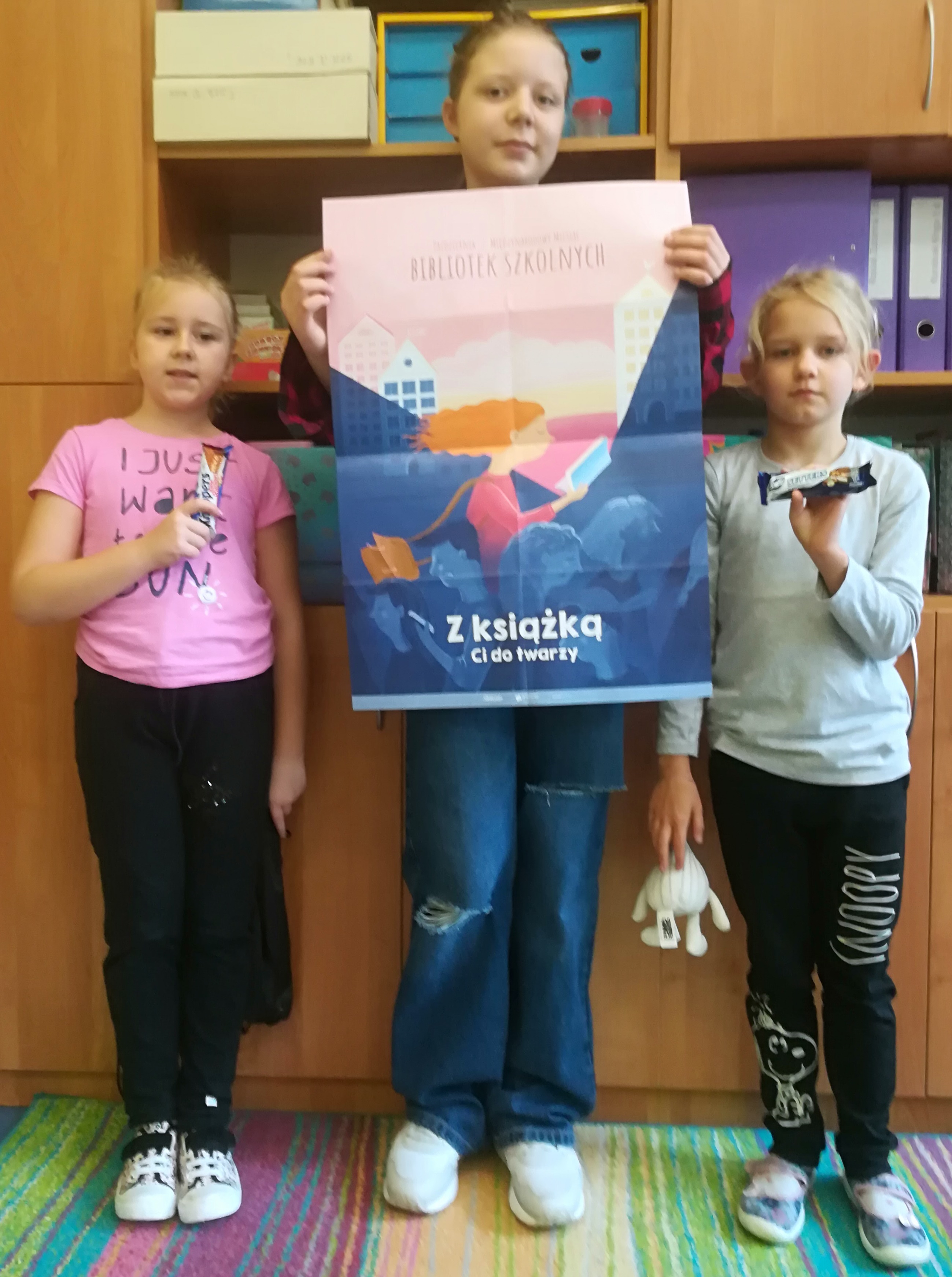 Trzy uczennice, jedna z nich trzyma w rękach plakat zatytułowany "Październik 2022 miesiącem bibliotek szkolnych".