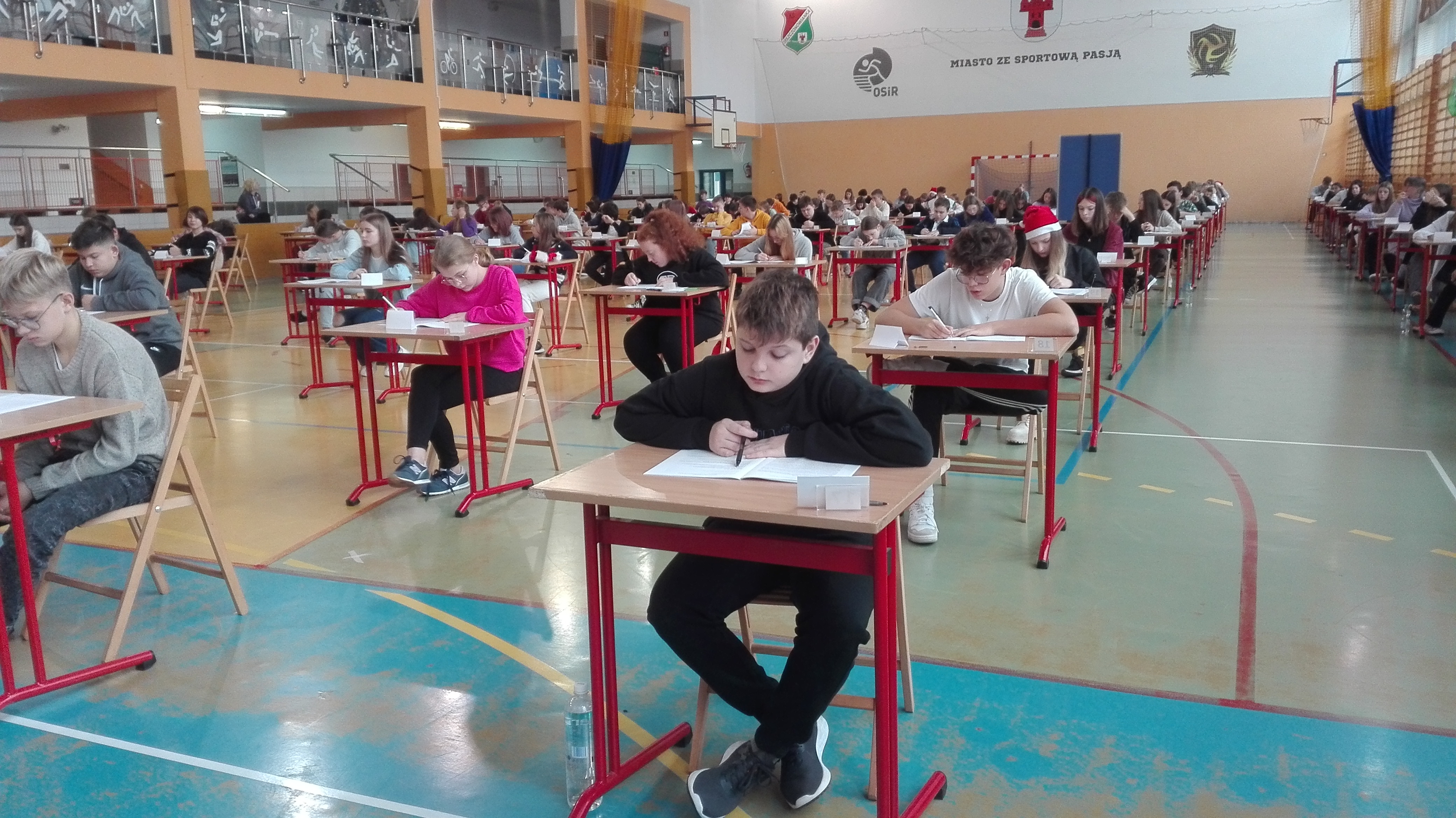 Uczniowie siedzący w pojedynczych ławkach na hali sportowej i piszący próbny egzamin.