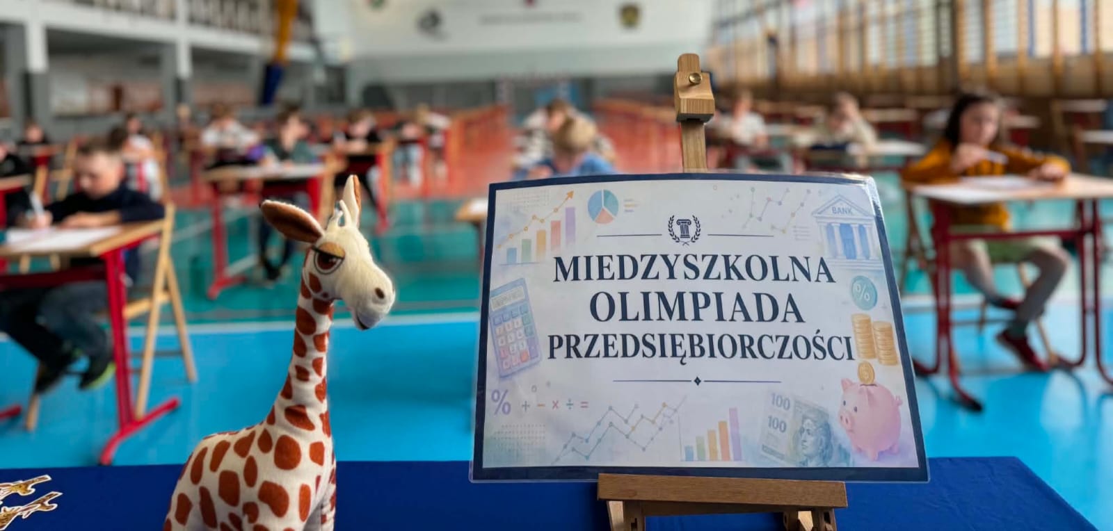 Plakat informacyjny na sztaludze, obok maskotka żyrafy, w tle uczniowie piszący test.